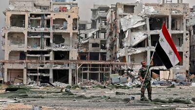 A member of the Syrian pro-government forces carries the national flag as he stands in front of damaged buildings in the Yarmouk Palestinian refugee camp on the southern outskirts of the capital Damascus on May 22, 2018. Louai Beshara / AFP