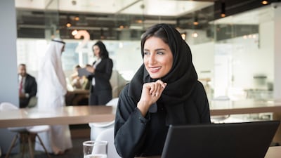 A photo of smiling Emirati businesswoman with laptop. Middle Eastern professional in traditional arab attire of abaya and hijab. She is sitting at conference table. Office worker is looking away while working in brightly lit office.Dubai, United Arab Emirates.