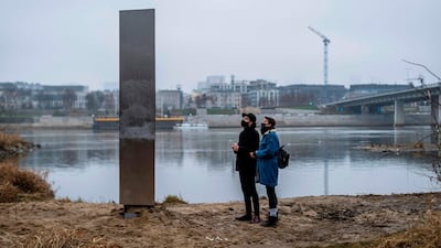 People look on December 10, 2020 at a metal monolith that has popped up on a riverbank of the Vistula in the Polish capital Warsaw, the latest in a string of similar objects that have recently appeared in Europe and the US. Similar-looking objects were found in the United States, Romania and Britain. / AFP / Wojtek RADWANSKI