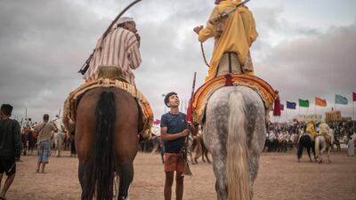 In this Thursday, July 25, 2019 photo, a boy hands a rifle to horse riders waiting for their turn to take part in an equestrian show known as Fantasia or Tabourida, in the coastal town of El Jadida, Morocco.