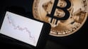 This illustration photograph shows a phone with a downward trend in the stock market in front of a screen displaying a Bitcoin logo, in Mulhouse on December 5, 2025. (Photo by SEBASTIEN BOZON / AFP)