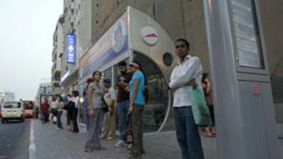 People wait to board a bus outside a non-working air-conditioned bus shelter, where the inside temperature is warmer than on the street.