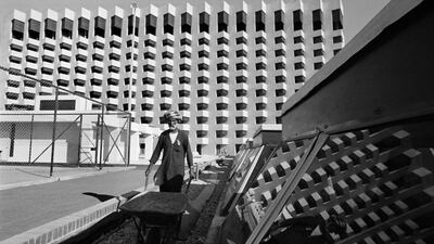 An expatriate worker pushes a wheel barrow past a modern apartment block. Abu Dhabi, 1975.